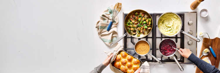 An overhead view shows a white kitchen counter and a gas stovetop with four burners. Two Misen stainless pans on the back burners hold roasted Brussels sprouts and mashed potatoes with chives. Two Misen Sauciers on the front burners contain gravy and cranberry sauce. Hands are stirring the cranberry sauce and holding a Misen Roasting Pan of dinner rolls. A green striped Misen Kitchen Towel with a whisk and a wooden cutting board with butter and a Misen Serrated Chef Spoon and Masher is next to the stovetop.