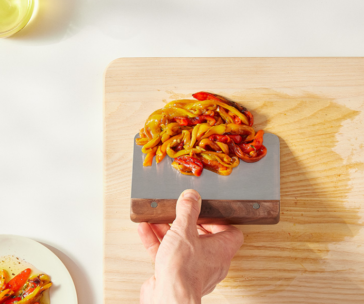 Using a walnut Misen bench scraper to scoop up sauteéed bell peppers off of a wooden cutting board