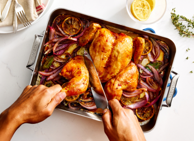 An overhead shot of a person carving a roasted chicken in a Misen Stainless Steel Roasting Pan. The chicken is surrounded by roasted red onions and lemon slices. The roasting pan has the 