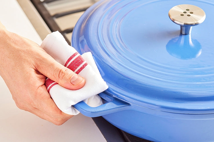 A close view of one of a blue Misen Enameled Cast Iron Braiser’s wide handle, as a hand easily grips it with a kitchen towel.