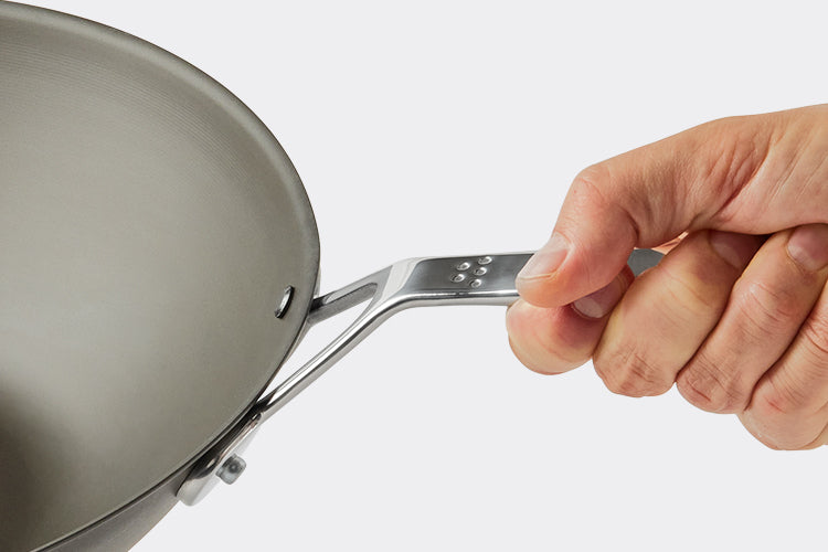 A hand holds the handle of a Misen Carbon Steel Wok on a white background. Half of the wok is visible.
