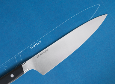 The image shows a Misen 8 inch Chef's Knife 2.0 with a black handle and a silver ACUTO440 steel blade on a blue background. The knife is positioned horizontally, with its handle on the left. Above the knife, a faint outline of the same knife is visible, suggesting a blueprint. The word 