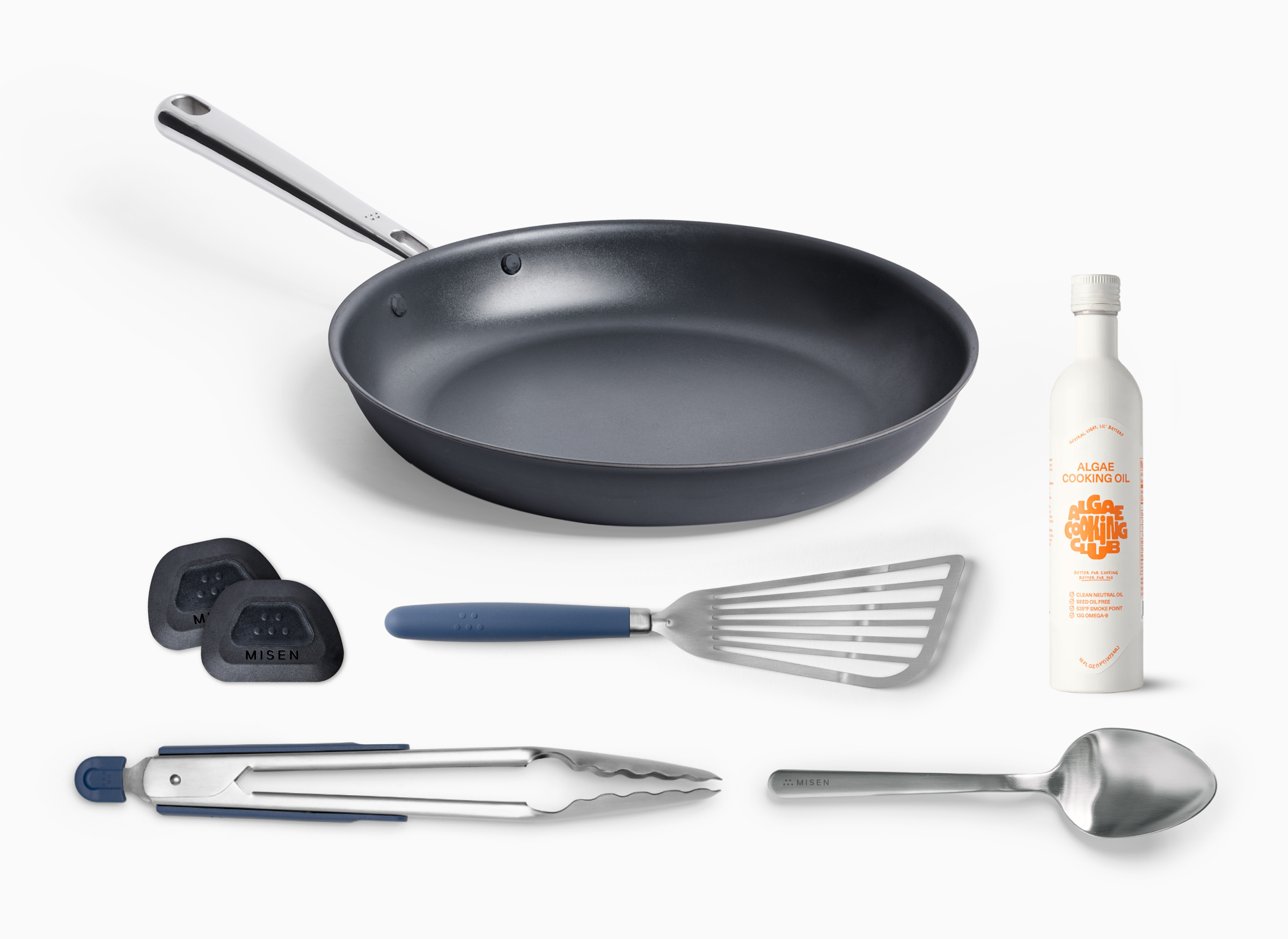 A top-down shot of a 10-inch Misen Carbon Nonstick™ Frying Pan. Next to the pan are two black Misen Pan Scrapers, a blue and silver slotted spatula, a pair of 13 inch Misen Stainless Steel Tongs, and a Misen Plating Chef Spoon There is also a 16 oz bottle of Algae Cooking Club Cooking Oil.