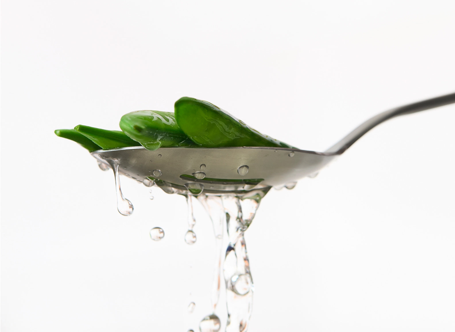 A stainless steel slotted spoon holds several green snap peas, with water dripping from the slots and the underside of the spoon. The spoon is angled slightly upwards, with the handle extending towards the right, against a plain white background.