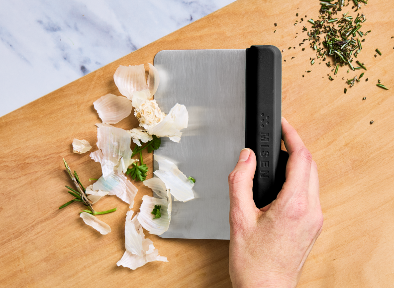 A person's right hand holds a Misen Bench Scraper with a black handle, pushing a pile of garlic skin, parsley, and rosemary across a light wooden cutting board. A small pile of chopped herbs is visible in the upper right corner.