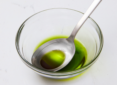 A close-up view shows a stainless steel Misen Measuring Chef Spoon resting in a clear glass bowl containing a green olive oil. The spoon is etched with measurements for 1 tbsp and 1 tsp.